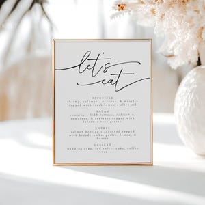 May include: A white menu with a gold frame and the text "let's eat" in black cursive. The menu lists appetizers, salad, entree, and dessert options.