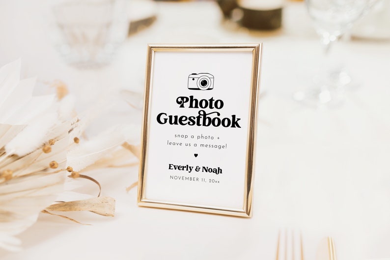 May include: A gold-framed sign with a white background and black text that reads "Photo Guestbook" with a camera icon. The text below reads "snap a photo + leave us a message!" and "Everly & Noah" with the date "NOVEMBER 11, 20xx".