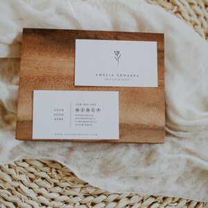 May include: A white business card with a simple floral design and the text "Amelia Edwards Photography". Below it is a second white business card with the text "Your Logo Here", a phone number, social media icons, and the text "www.yourwebsite.com". Both cards are on a wooden surface.