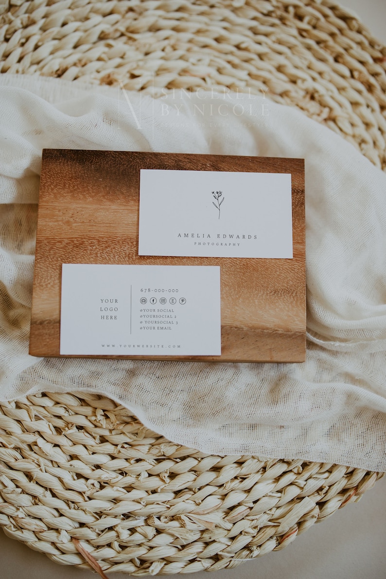 May include: A white business card with a simple floral design and the text "Amelia Edwards Photography". Below it is a second white business card with the text "Your Logo Here", a phone number, social media icons, and the text "www.yourwebsite.com". Both cards are on a wooden surface.