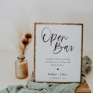 May include: A white sign with gold trim and the words "Open Bar" in elegant script. The sign includes the text "tonight's drinks are free & tomorrow's stories will be priceless" and a couple's names and date. A decorative vase and fabric are in the background.