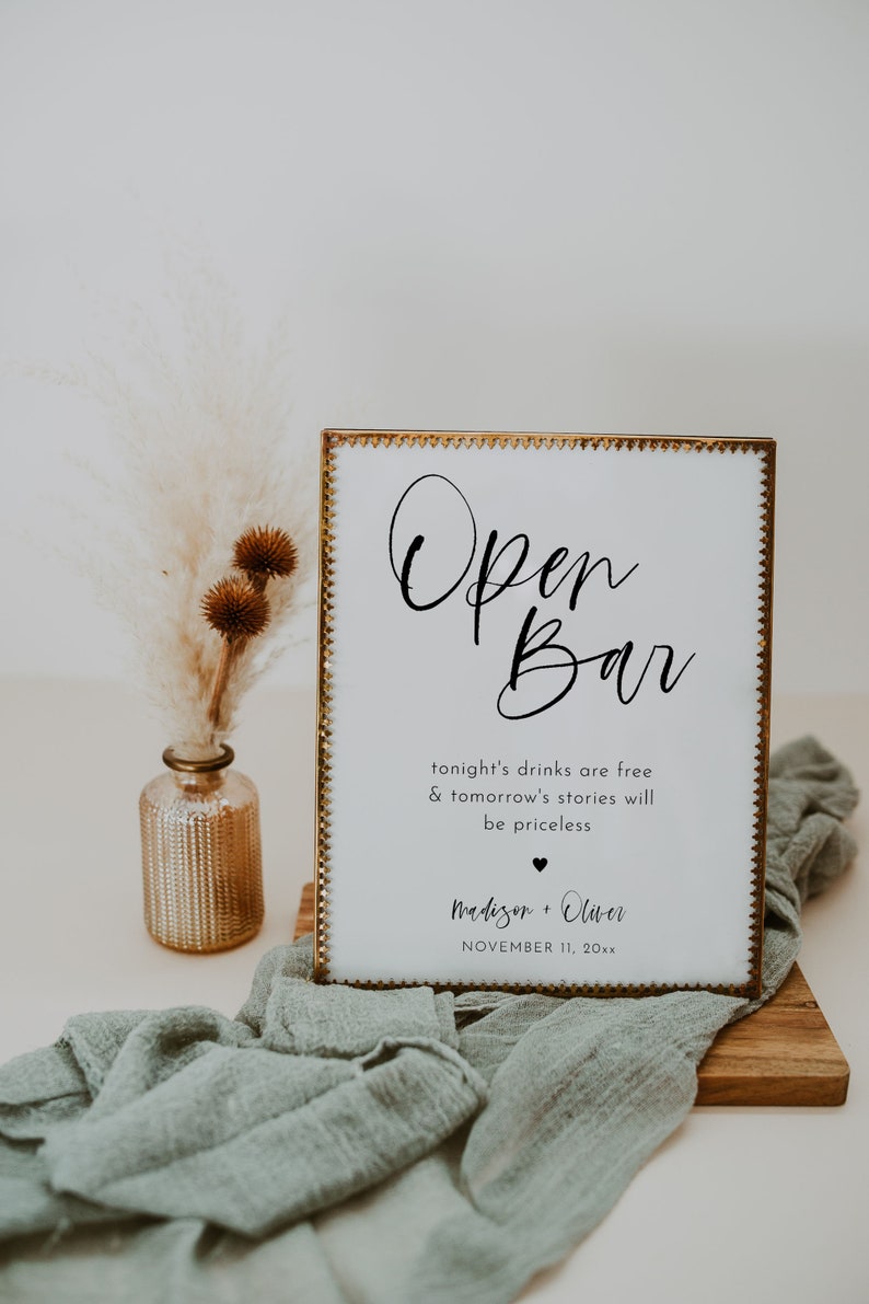May include: A white sign with gold trim and the words "Open Bar" in elegant script. The sign includes the text "tonight's drinks are free & tomorrow's stories will be priceless" and a couple's names and date. A decorative vase and fabric are in the background.