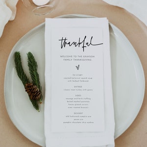 May include: A white card with black text on a white plate. The text reads "thankful" and lists the menu for a Thanksgiving dinner. The menu includes roasted butternut squash soup, classic roast turkey with gravy, sausage and herb stuffing, baked mashed potatoes, honey glazed carrots, oven roasted broccoli, old fashioned pumpkin pie, pecan pie, and pumpkin chocolate chip cookies.