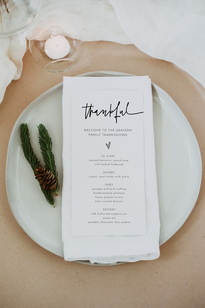 May include: A white card with black text on a white plate. The text reads "thankful" and lists the menu for a Thanksgiving dinner. The menu includes roasted butternut squash soup, classic roast turkey with gravy, sausage and herb stuffing, baked mashed potatoes, honey glazed carrots, oven roasted broccoli, old fashioned pumpkin pie, pecan pie, and pumpkin chocolate chip cookies.