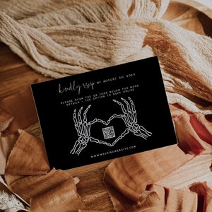 Elegant black RSVP card featuring skeleton hand graphics forming a heart, with a QR code for online response. Set against a warm, textured fabric background. Customizable with wedding details and website link.