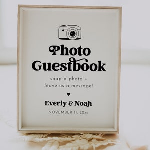 May include: A white sign with a black camera icon and the text "Photo Guestbook" in a retro font. The sign also includes the text "snap a photo + leave us a message!" and the names "Everly & Noah" with the date "NOVEMBER 11, 20xx".