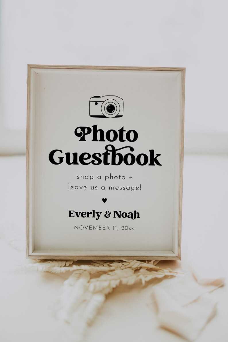 May include: A white sign with a black camera icon and the text "Photo Guestbook" in a retro font. The sign also includes the text "snap a photo + leave us a message!" and the names "Everly & Noah" with the date "NOVEMBER 11, 20xx".