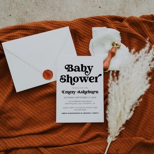 Op de afbeelding: Een witte babyshower-uitnodiging met zwarte tekst. De uitnodiging luidt "Baby Shower" in een retro-lettertype, gevolgd door "honoring mom-to-be Emery Asheburn". De datum, locatie en RSVP-informatie staan hieronder vermeld.