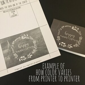 以下が含まれることがあります： 白い花輪と「happy anniversary」という筆記体で書かれたテキストが印刷されたカードのクローズアップ。カードはグレーの背景に印刷されています。画像は、プリンターによって色がどのように異なるかを示しています。