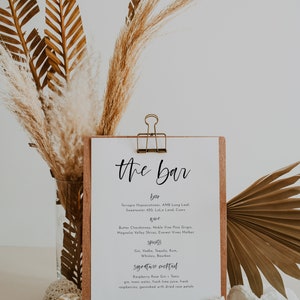 May include: A wooden bar menu sign with the words "the bar" in elegant script. The menu lists beer, wine, spirits, and a signature cocktail. Dried palm leaves and pampas grass decorate the background.