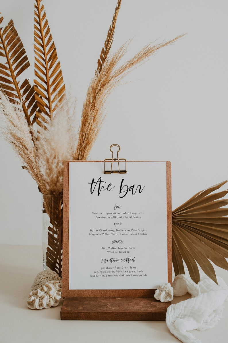 May include: A wooden bar menu sign with the words "the bar" in elegant script. The menu lists beer, wine, spirits, and a signature cocktail. Dried palm leaves and pampas grass decorate the background.