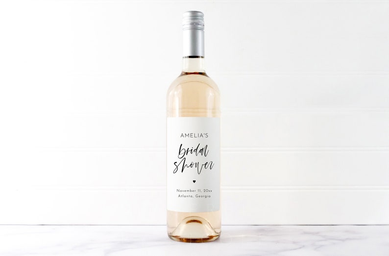 Może przedstawiać: Przezroczysta szklana butelka wina ze srebrną zakrętką i białą etykietą. Etykieta głosi "AMELIA'S Bridal Shower" elegancką czcionką, z datą i lokalizacją poniżej. Butelka zawiera jasno zabarwioną ciecz.