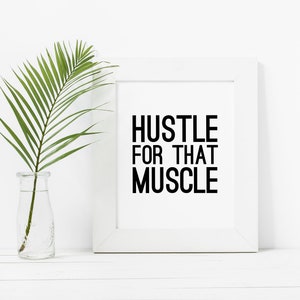 May include: White framed motivational poster with the text "Hustle for that Muscle" in black lettering. A green palm frond is in a clear glass vase on a white surface.