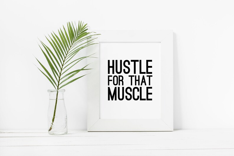 May include: White framed motivational poster with the text "Hustle for that Muscle" in black lettering. A green palm frond is in a clear glass vase on a white surface.
