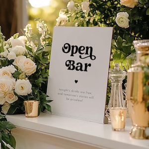 Op de afbeelding: Een wit bord met zwarte tekst die "Open Bar" in een retro-lettertype luidt. Onder de tekst staat een klein zwart hartje en de tekst "tonight's drinks are free, and tomorrow's stories will be priceless!"