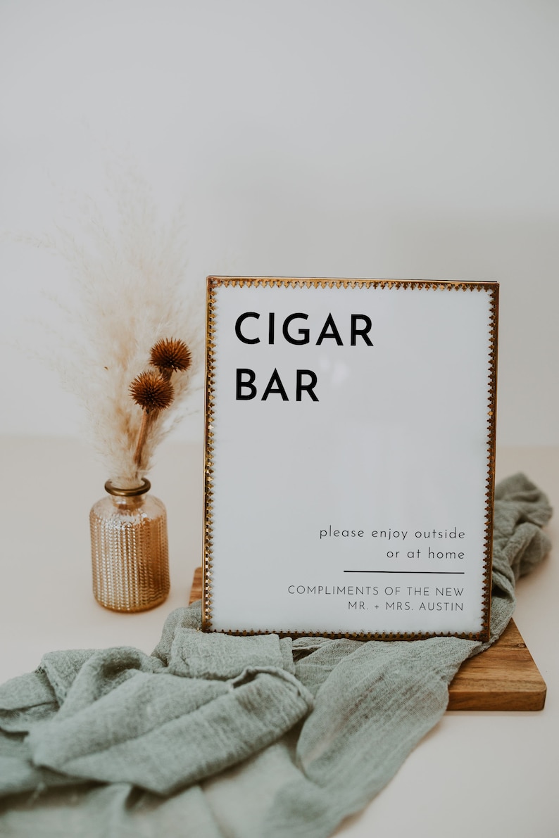 Puede incluir: Un cartel con marco dorado con el texto "CIGAR BAR" en letras negras. El cartel tambi&eacute;n incluye el texto "please enjoy outside or at home COMPLIMENTS OF THE NEW MR. + MRS. AUSTIN".