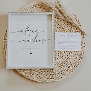 May include: A framed white sign with the words "advice + wishes" in elegant black script, with a small heart below. A matching card is next to the sign. The sign and card are on a woven circular mat, with dried decorative plants.