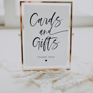 May include: A gold-framed sign with the words "Cards and Gifts" in elegant black script. Below, the text reads "Thank You!" with a small heart symbol. The sign is displayed on a white surface.