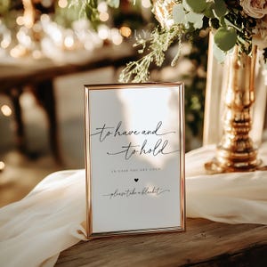 May include: A gold-framed sign with the text "to have and to hold, in case you get cold, please take a blanket" in black calligraphy. The sign is sitting on a white tablecloth.