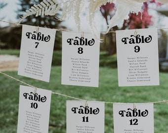May include: White cards with black text listing table numbers 7 through 12 and guest names. The cards are hanging from a string with a white fluffy plant and a white cloth hanging above.