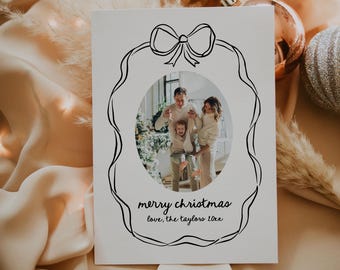 May include: A white Christmas card with a black and white illustration of a wreath with a bow. The wreath has a photo of a family of three inside. The text "merry christmas love, the taylors 10xx" is written below the wreath.