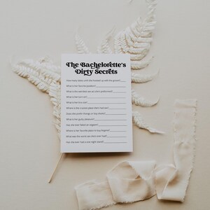 A bachelorette party game card titled "The Bachelorette’s Dirty Secrets" on a neutral background with white ferns and beige fabric. A fun and playful icebreaker featuring personal and cheeky questions.