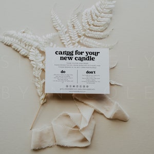 May include: A white card with black text that reads "caring for your new candle" with instructions on how to care for a candle. The card is on a light beige background with white dried ferns and a white ribbon.