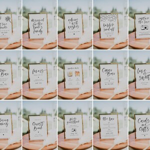 May include: A collection of framed wedding signs with gold frames and white backgrounds. Each sign features elegant black calligraphy with phrases like "Open Bar," "Capture the Love," and "Cards and Gifts." The signs are arranged on a wooden surface.