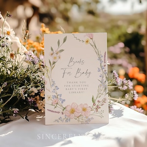 Books for baby sign featuring pastel wildflowers in a delicate wreath design with script and serif fonts, placed outdoors on a white table surrounded by blooming garden flowers. Perfect for a spring or summer baby shower with a soft floral theme.