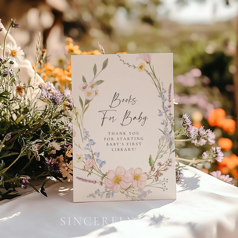Books for baby sign featuring pastel wildflowers in a delicate wreath design with script and serif fonts, placed outdoors on a white table surrounded by blooming garden flowers. Perfect for a spring or summer baby shower with a soft floral theme.