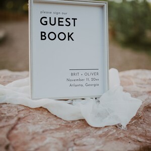 Puede incluir: Un cartel blanco con texto negro que dice "Por favor, firme nuestro LIBRO DE VISITAS" con los nombres Brit + Oliver, 11 de noviembre de 20xx, Atlanta, Georgia debajo.