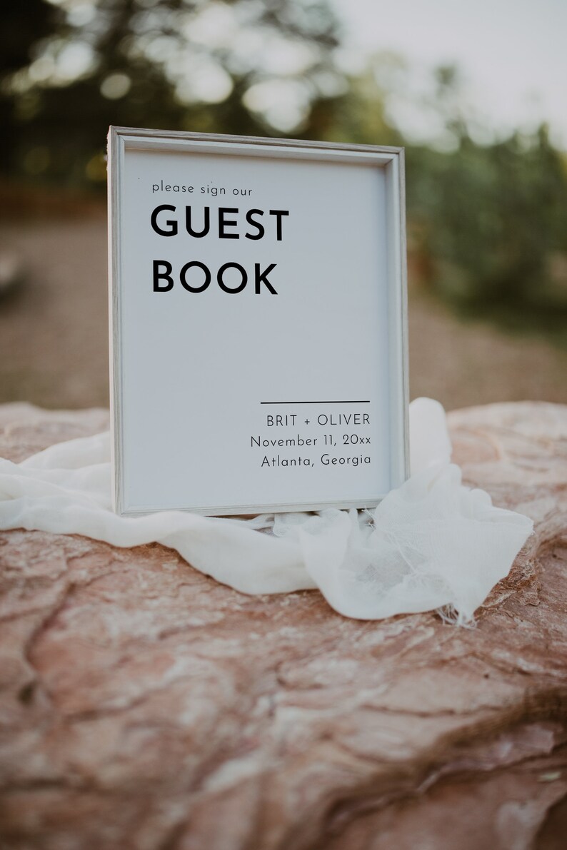 Puede incluir: Un cartel blanco con texto negro que dice "Por favor, firme nuestro LIBRO DE VISITAS" con los nombres Brit + Oliver, 11 de noviembre de 20xx, Atlanta, Georgia debajo.