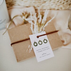 May include: A wrapped gift box with a brown paper exterior, tied with a dark brown cord. A white tag with three green wreath illustrations and the text "merry christmas + happy new year" is attached. Dried flowers and a woven basket are in the background.