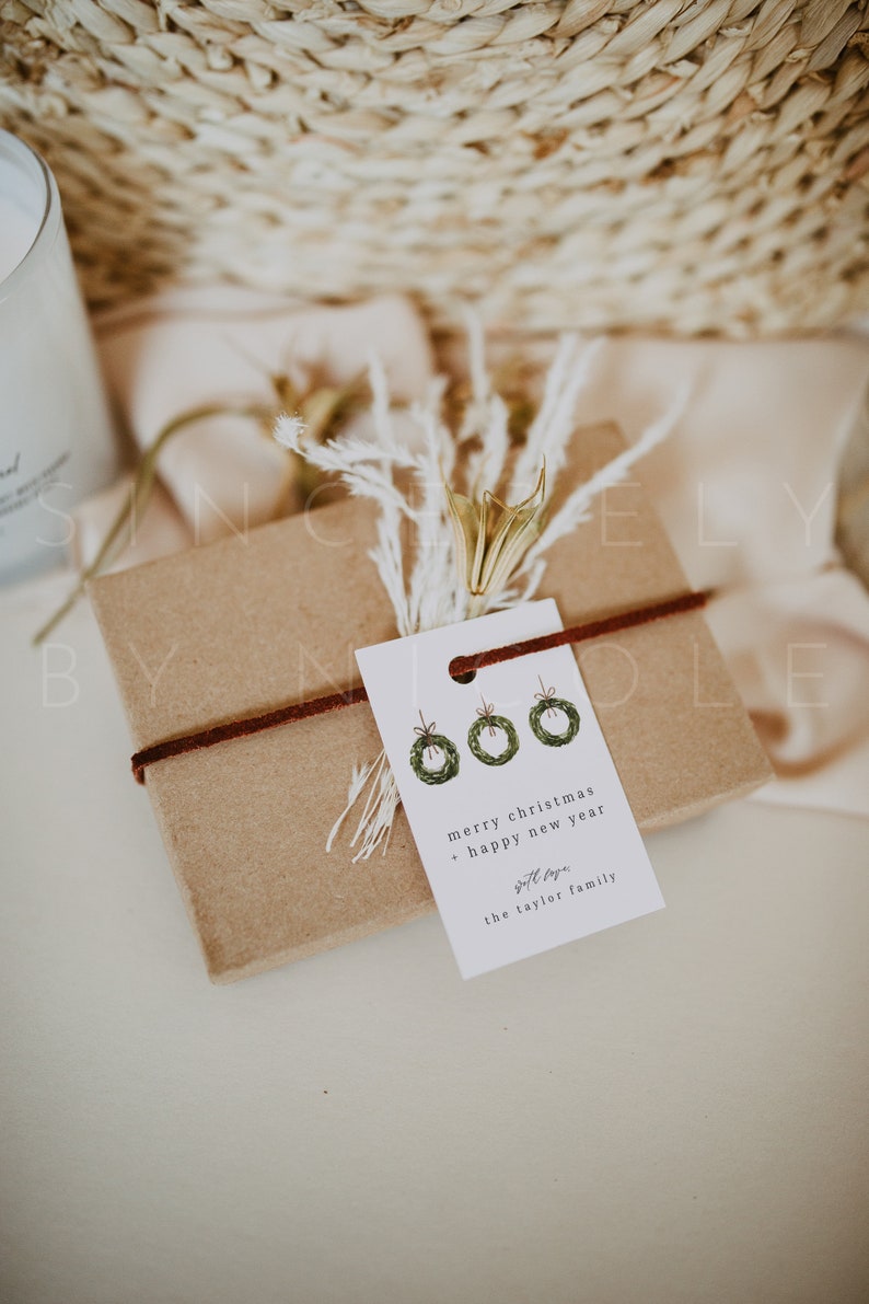 May include: A wrapped gift box with a brown paper exterior, tied with a dark brown cord. A white tag with three green wreath illustrations and the text "merry christmas + happy new year" is attached. Dried flowers and a woven basket are in the background.
