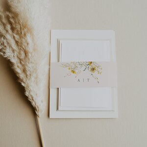 May include: A white invitation suite with a floral design and the text "A | T" on the band. The invitation is on a beige background with a dried pampas grass.