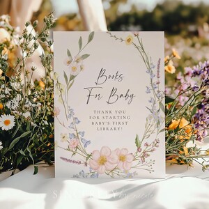 Delicate floral baby shower book request sign framed by soft wildflowers, displayed among daisies and greenery for a natural garden setting. A charming way to invite guests to build baby’s first library.