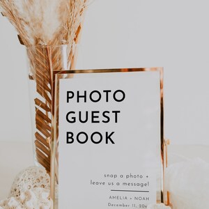 Puede incluir: Un cartel con marco dorado con el texto "PHOTO GUEST BOOK" y las instrucciones "snap a photo + leave us a message!"  El cartel tambi&eacute;n incluye los nombres "AMELIA + NOAH" y la fecha "December 11, 20x".