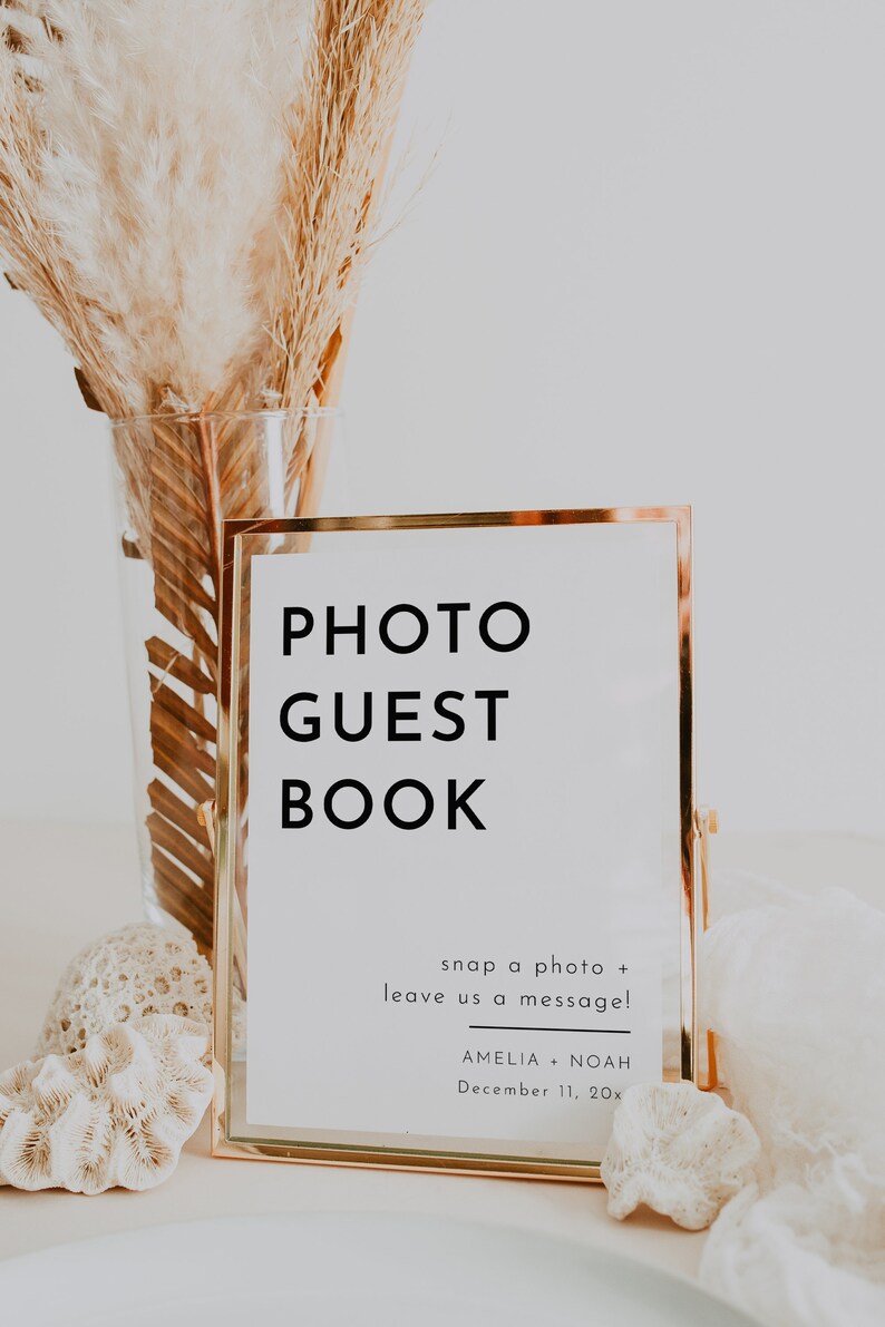 Puede incluir: Un cartel con marco dorado con el texto "PHOTO GUEST BOOK" y las instrucciones "snap a photo + leave us a message!"  El cartel tambi&eacute;n incluye los nombres "AMELIA + NOAH" y la fecha "December 11, 20x".