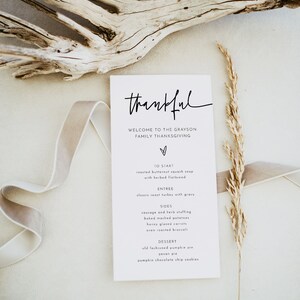 May include: A white card with black text that reads "thankful" and "Welcome to the Grayson Family Thanksgiving". The card lists the menu for a Thanksgiving dinner, including roasted butternut squash soup, classic roast turkey with gravy, sausage and herb stuffing, baked mashed potatoes, honey glazed carrots, oven roasted broccoli, old fashioned pumpkin pie, pecan pie, and pumpkin chocolate chip cookies.