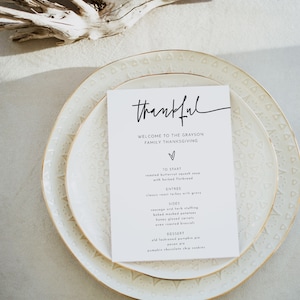 May include: A white card with black text on a white plate with a gold rim. The card says "thankful" and lists the menu for a Thanksgiving dinner. The menu includes roasted butternut squash soup, classic roast turkey with gravy, sausage and herb stuffing, baked mashed potatoes, honey glazed carrots, oven roasted broccoli, old fashioned pumpkin pie, pecan pie, and pumpkin chocolate chip cookies.