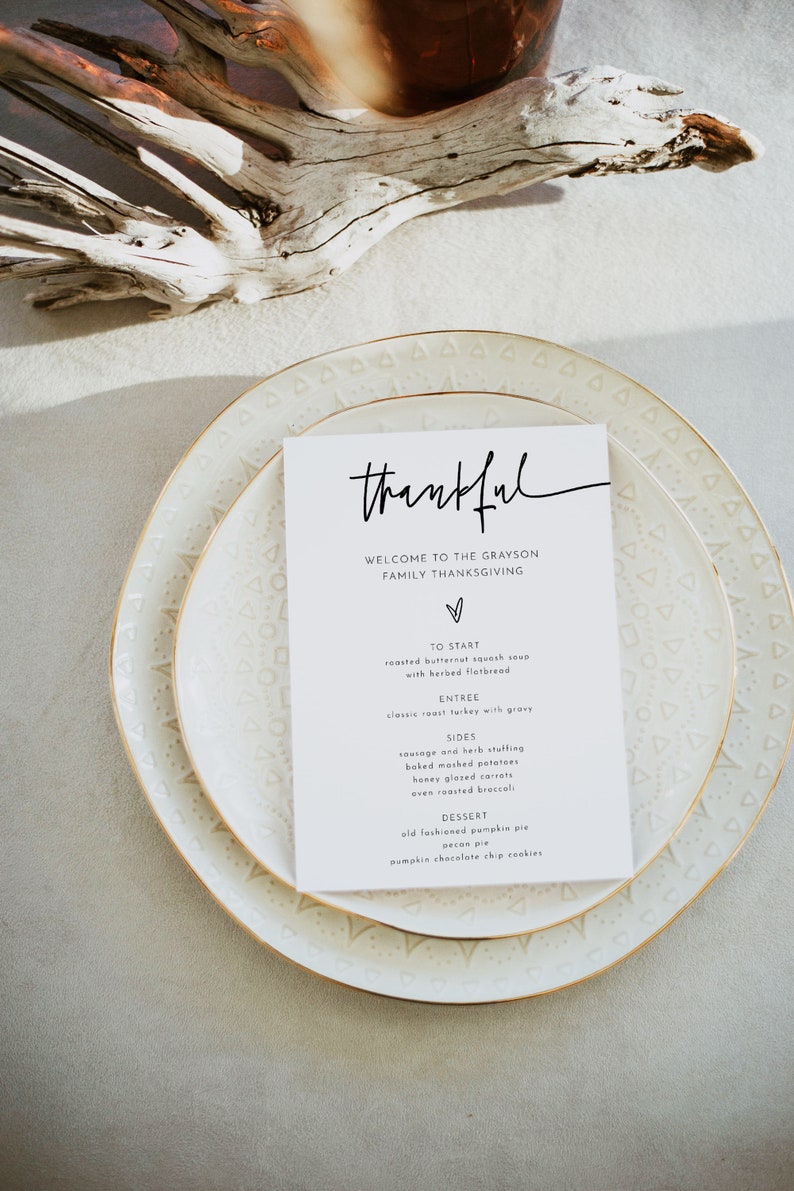 May include: A white card with black text on a white plate with a gold rim. The card says "thankful" and lists the menu for a Thanksgiving dinner. The menu includes roasted butternut squash soup, classic roast turkey with gravy, sausage and herb stuffing, baked mashed potatoes, honey glazed carrots, oven roasted broccoli, old fashioned pumpkin pie, pecan pie, and pumpkin chocolate chip cookies.
