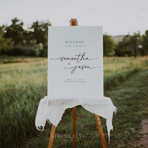 Wedding welcome sign on an easel with soft fabric, set in an open field with lush greenery. A simple yet elegant touch for outdoor weddings and nature-inspired themes.