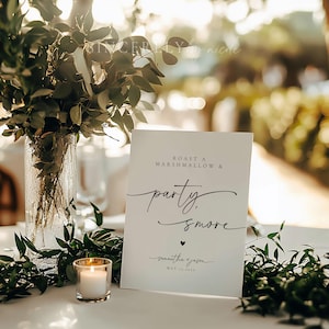 May include: White sign with black text that reads "Roast a Marshmallow & party. smore" with a small heart and the text "Samantha & Jason May 11, 2023" below. The sign is sitting on a table with greenery and a candle.