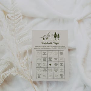 A bridal party bingo game card placed on plush white fabric, accented with delicate dried florals. The design features a nature-inspired aesthetic with elegant typography, making it a stylish addition to bachelorette festivities.