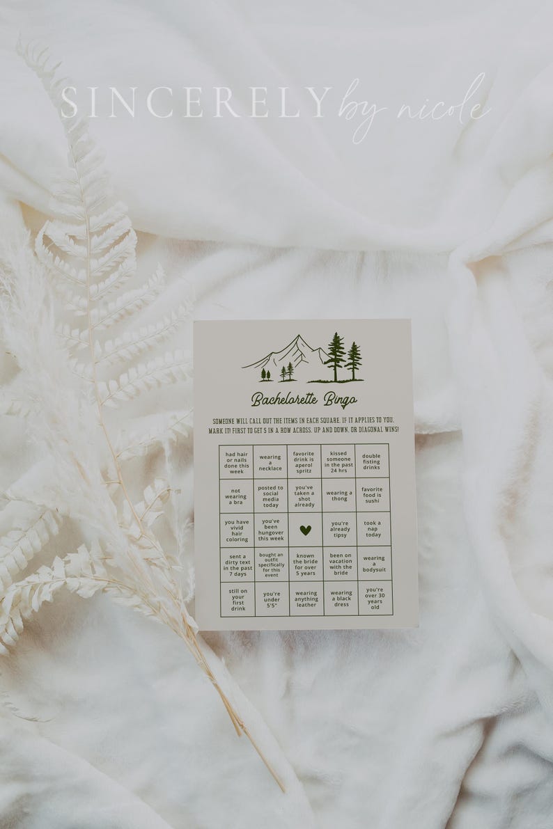 A bridal party bingo game card placed on plush white fabric, accented with delicate dried florals. The design features a nature-inspired aesthetic with elegant typography, making it a stylish addition to bachelorette festivities.