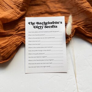 A printed bachelorette party game titled "The Bachelorette’s Dirty Secrets" placed on a wooden surface with neutral fabric and a dried palm leaf. A fun way to add laughter and spice to the celebration.