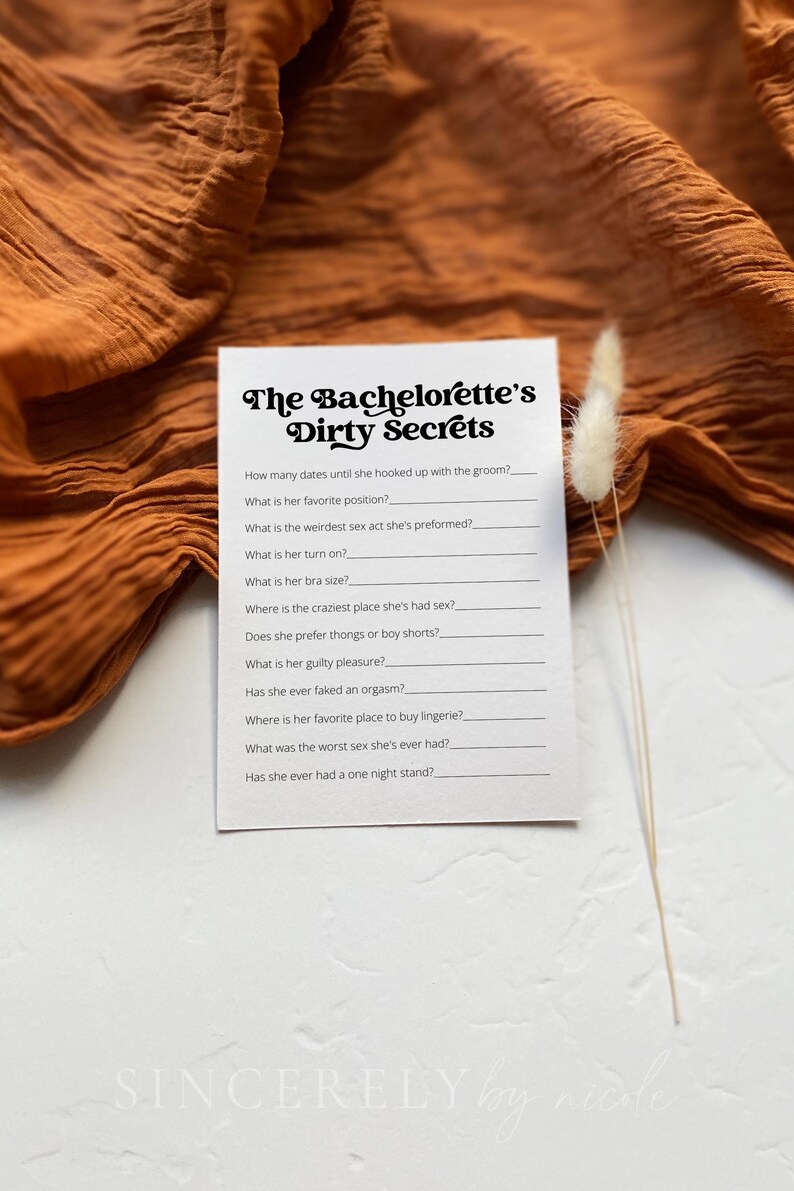 A printed bachelorette party game titled "The Bachelorette’s Dirty Secrets" placed on a wooden surface with neutral fabric and a dried palm leaf. A fun way to add laughter and spice to the celebration.
