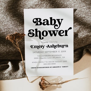 Op de afbeelding: Een witte baby shower uitnodiging met zwarte tekst. De tekst luidt "Baby Shower" in een retro lettertype, gevolgd door "honoring mom-to-be Emery Asheburn". De datum, locatie en RSVP-informatie zijn ook inbegrepen.