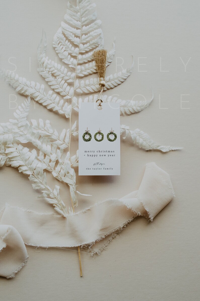 May include: A white gift tag with three green wreath illustrations, hanging from a gold string. The tag reads "merry christmas + happy new year" and includes a family name. The tag is set against a backdrop of white fern and cream-colored ribbon.