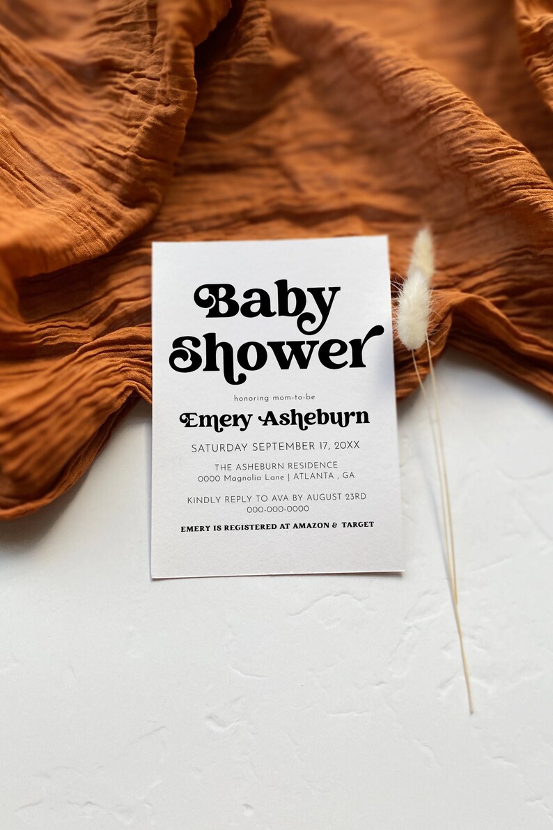 Op de afbeelding: Een witte babyborrel-uitnodiging met zwarte tekst. De uitnodiging luidt "Baby Shower" in een retro-lettertype, gevolgd door "honoring mom-to-be Emery Asheburn". De datum, tijd en locatie van de borrel staan hieronder vermeld. De uitnodiging bevat ook een telefoonnummer en een notitie dat Emery is geregistreerd bij Amazon en Target.